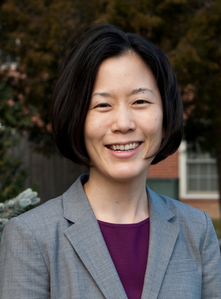 Person wearing a light gray blazer over a dark purple top, standing outdoors with trees and a brick building in the background