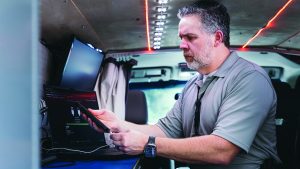 The image shows a man seated inside a vehicle, possibly a van, which appears to be outfitted for technical or surveillance work. He is wearing a gray polo shirt and has short hair. The man is holding a smartphone in his right hand and interacting with a laptop using his left hand. The laptop screen displays what looks like technical data or a software interface. There are LED lights on the ceiling of the vehicle, some of which are red and others white. In the background, there are various pieces of equipment including monitors and cables attached to the interior walls of the vehicle.