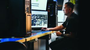 The image shows a person seated at a desk in front of multiple computer monitors. The individual is using a mouse with their right hand and appears to be focused on the screens. One of the monitors displays a detailed map or satellite image, while another screen shows what looks like data or graphs. The desk has various items including a keyboard and other electronic equipment. The environment suggests a high-tech workspace, possibly related to surveillance or data analysis.