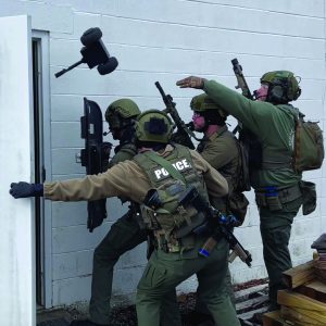 Four officers in tactical gear breach door; one uses battering ram, another deploys small robot; 'POLICE' visible on back; active entry in progress.