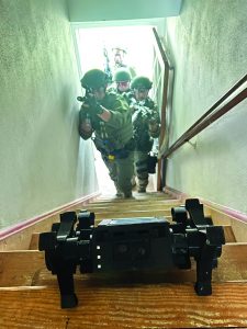 Four armed officers in tactical gear ascend wooden stairs with weapons drawn, focusing on a black device placed on steps ahead; active operation in progres