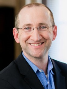 Person in a dark suit jacket over a light blue collared shirt with white stripes, standing indoors near a bright window and blurred background
