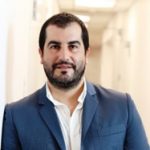 Person wearing a dark blue suit jacket over a white collared shirt, standing in a brightly lit hallway with white walls and doors in the background