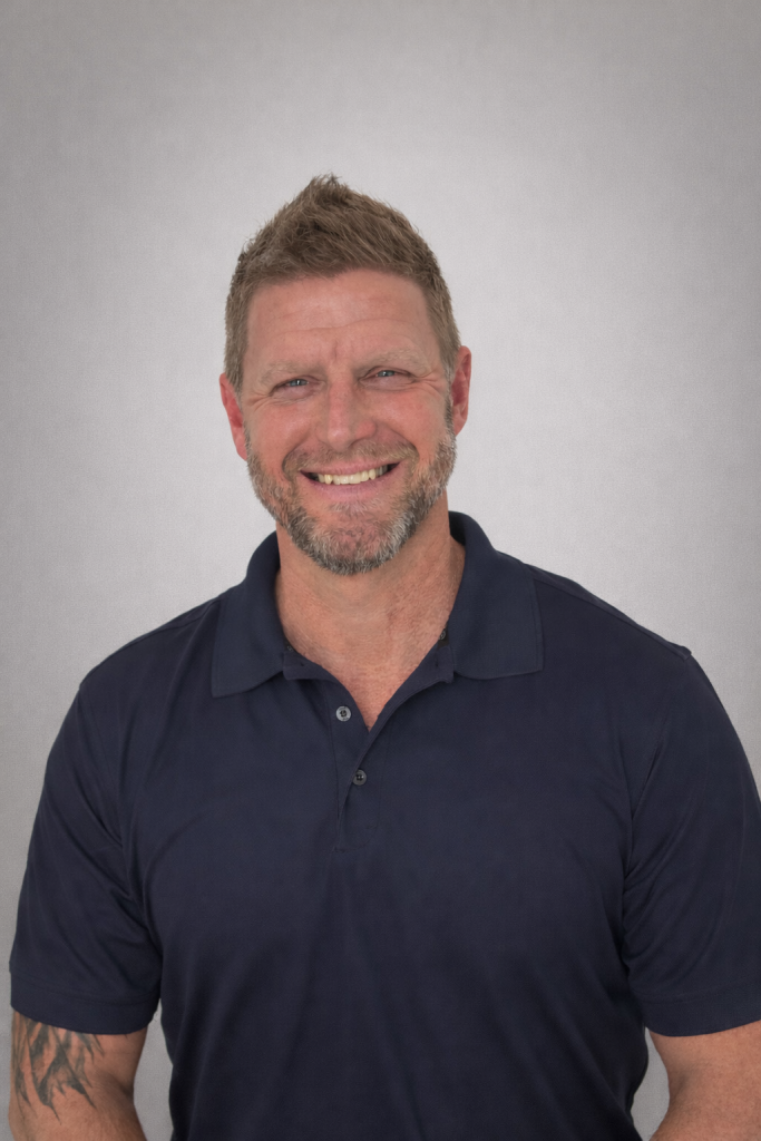 Studio portrait of an individual wearing a dark navy polo shirt, standing against a plain gray background, with short sleeves revealing a forearm tattoo and even, soft lighting