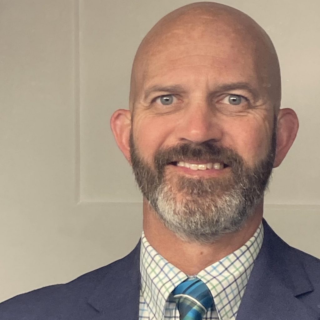 Individual in blue suit jacket, white-green checkered shirt, and blue striped tie stands before plain beige background