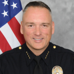 Uniformed officer stands before American flag; name tag reads 'STANSBROOK,' badge visible on right chest, emphasizing service and national pride