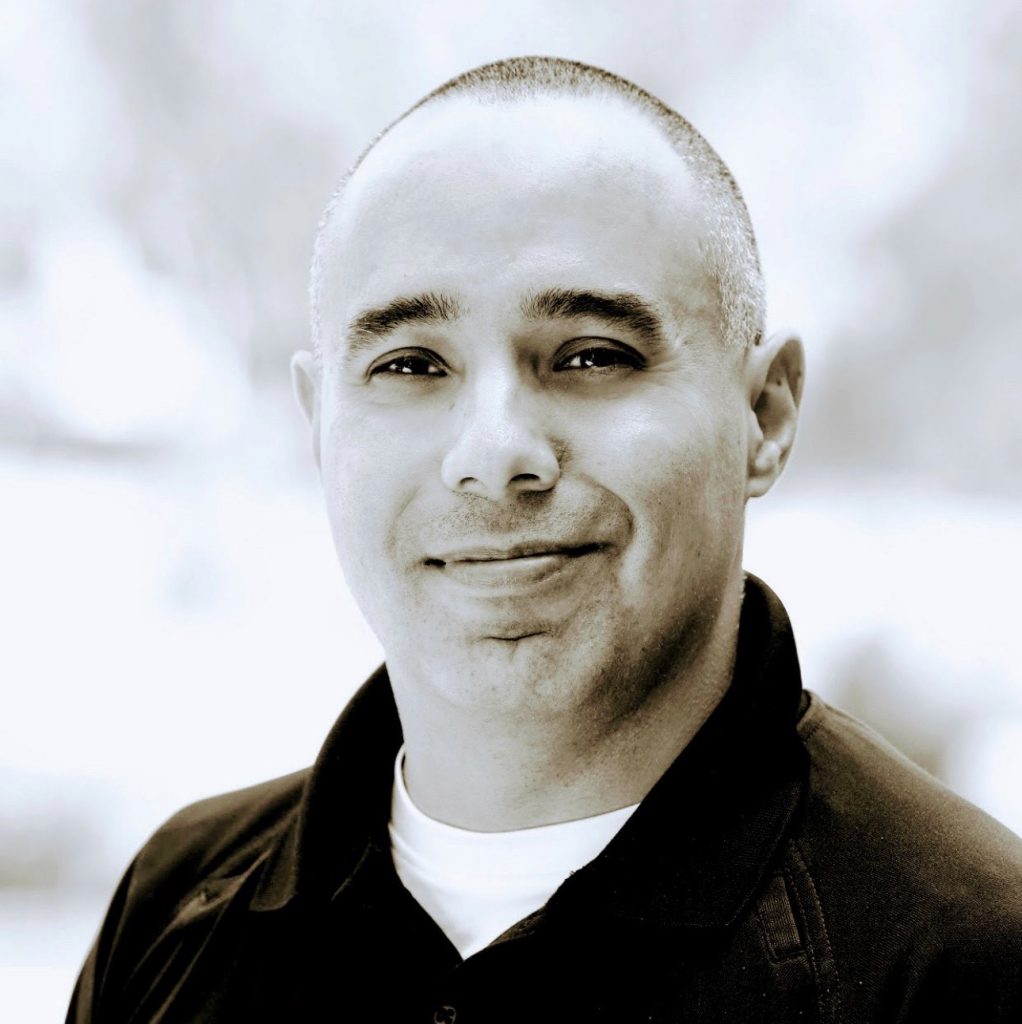 Person in black collared shirt over white undershirt; background softly blurred with mixed light and dark tones