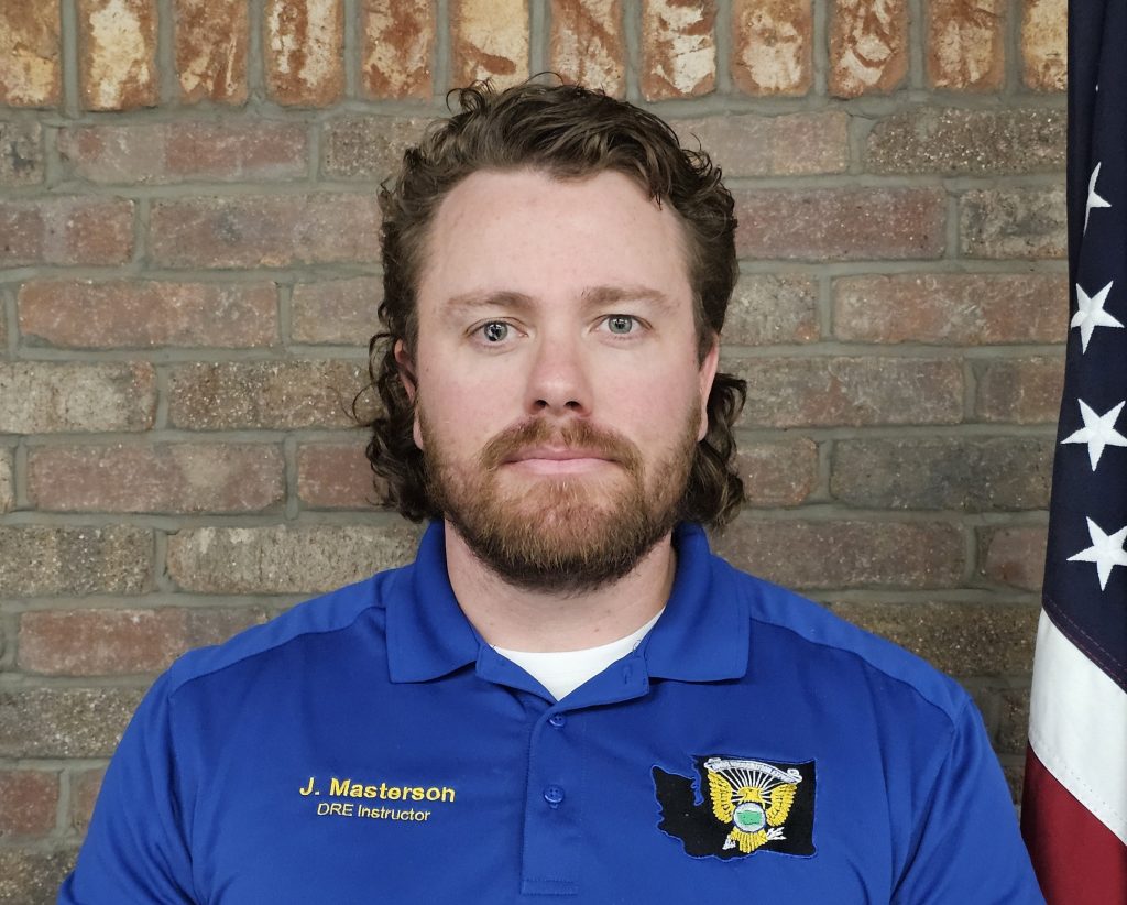 An individual wearing a blue polo with embroidered name and patch stands before a brick wall with a U.S. flag visible along the right edge positioned