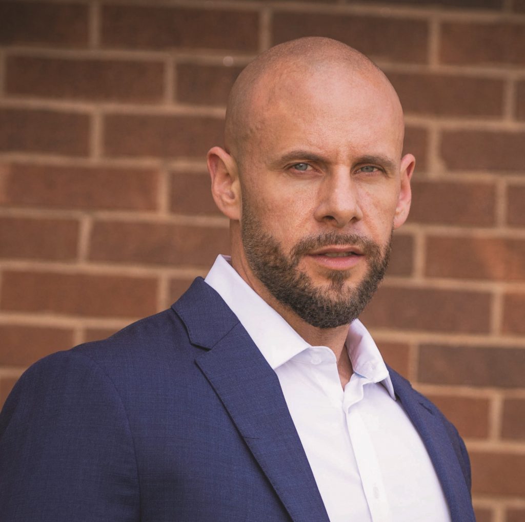 Person wearing a navy blue suit jacket over a crisp white dress shirt, standing in front of a brick wall with horizontal pattern