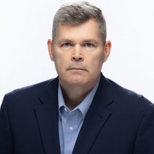 Individual with short gray hair wears dark navy blazer and light blue shirt against plain white background