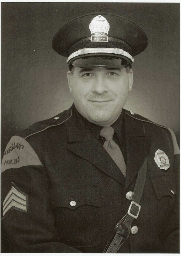 Black‑and‑white portrait of a uniformed Portland Police sergeant wearing peaked cap, tie, badge, service stripes, shoulder patch, and cross‑body strap against studio backdrop