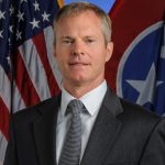 Man in suit and tie stands before U.S. and Tennessee flags against a blue backdrop; both flags are fully visible and prominently displayed behind him