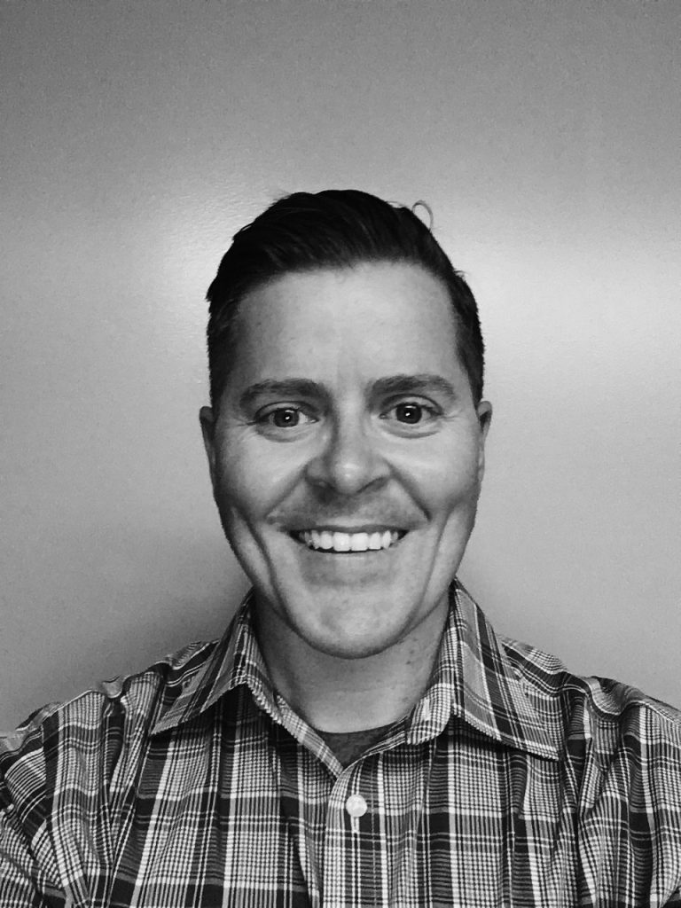 a black and white photograph of a person from the shoulders up. The person is wearing a plaid shirt with a collar, and the hair appears to be neatly combed back. The background is plain and without any distinct features or objects.