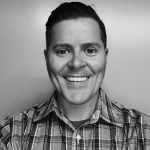 a black and white photograph of a person from the shoulders up. The person is wearing a plaid shirt with a collar, and the hair appears to be neatly combed back. The background is plain and without any distinct features or objects.