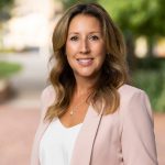 Person in light pink blazer and white top stands on brick pathway outdoors, surrounded by green trees and foliage; face is intentionally blurred