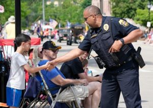 Uniformed police officer in short‑sleeve patrol uniform extends hand to greet a child along a parade route, with seated spectators, folding chairs, American flags, street barricades, and vehicles lining a city street.