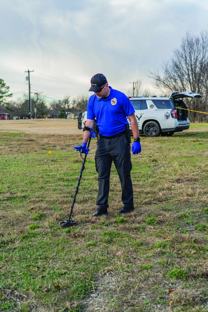An officer uses a handheld metal detector to scan grassy terrain marked with evidence flags, with a marked patrol SUV, open rear hatch, and crime scene tape behind