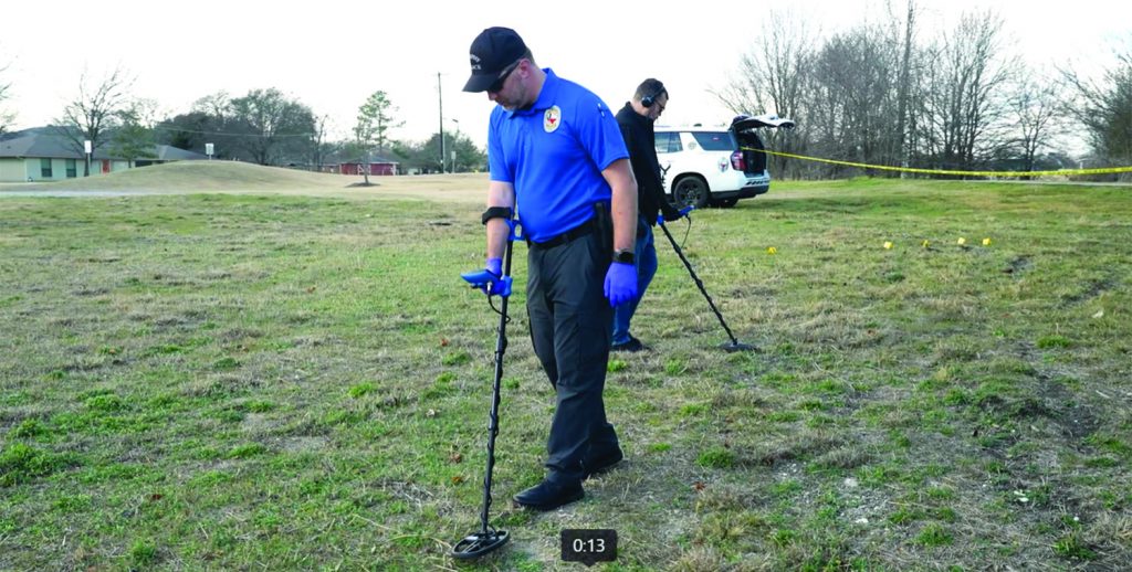Crime scene investigators search a grassy field using handheld metal detectors while wearing gloves, with evidence markers, caution tape, and a police SUV nearby