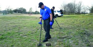 Crime scene investigators search a grassy field using handheld metal detectors while wearing gloves, with evidence markers, caution tape, and a police SUV nearby