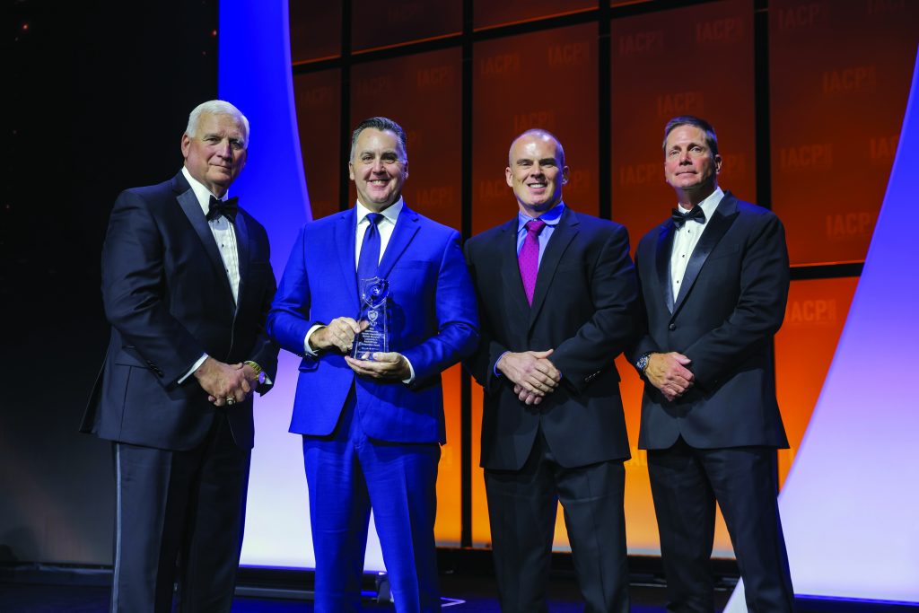 Four individuals in formal suits stand on stage with orange IACP backdrop; center person in blue suit holds a glass award under bright event lighting.