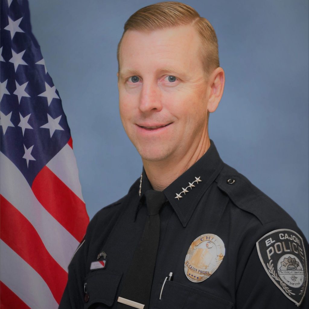 El Cajon police officer in uniform stands before U.S. flag; badge and shoulder patch clearly visible; and focus on attire