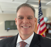 Individual in suit and tie stands in office with American flag, globe, and bookshelves in background;