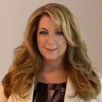 Individual with long wavy blonde hair in white blazer and black-and-white patterned blouse stands against plain beige background