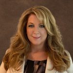 Person with long, voluminous, wavy blonde hair wearing a white blazer over a black patterned top, posed against a plain dark background