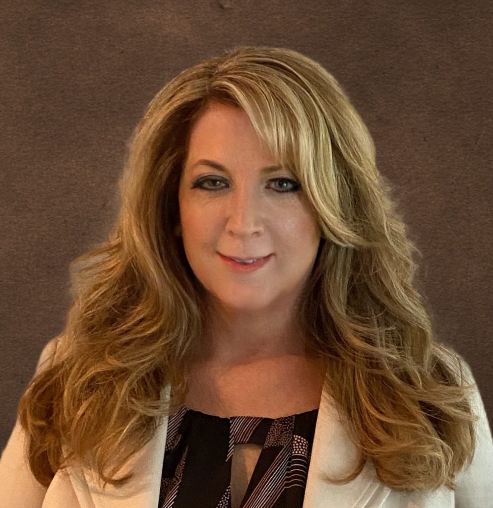 Person with long, voluminous, wavy blonde hair wearing a white blazer over a black patterned top, posed against a plain dark background