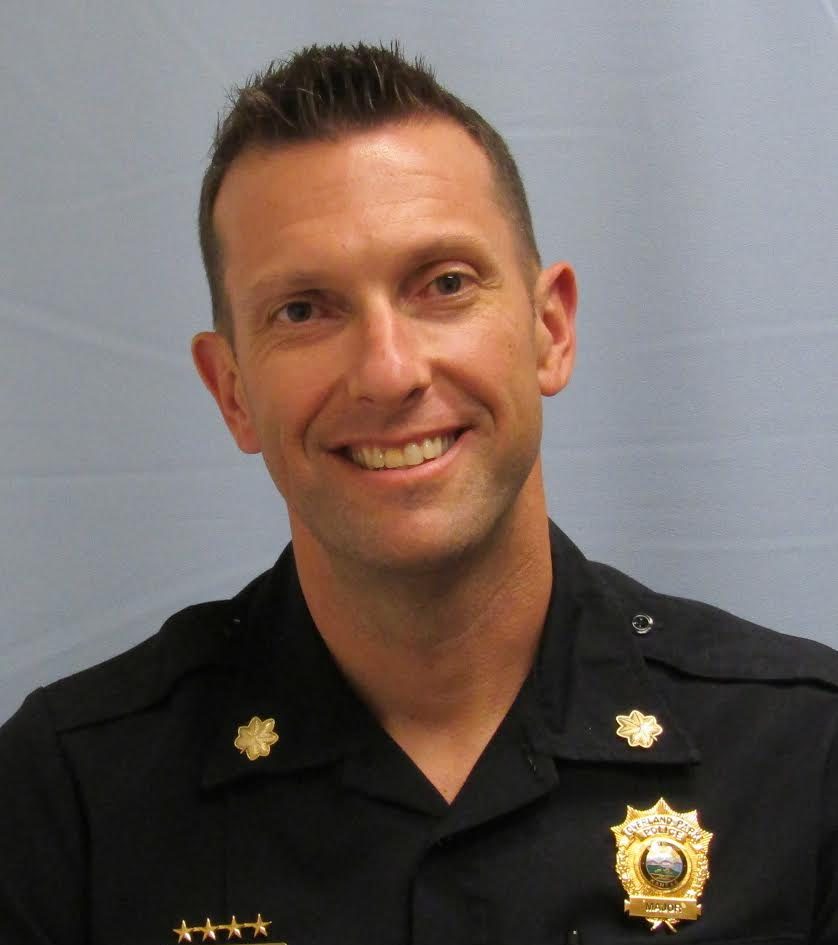 Officer in a dark uniform with gold oak‑leaf collar pins, four‑star nameplate, and a detailed badge stands before a light gray backdrop.