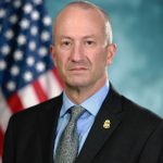 Individual in black suit jacket with gold badge, blue shirt, and patterned tie stands before American flag and blurred teal background