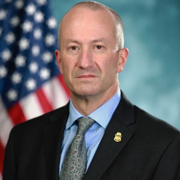Individual in black suit jacket with gold badge, blue shirt, and patterned tie stands before American flag and blurred teal background
