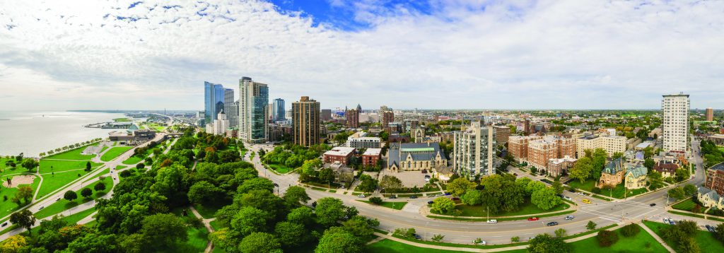 Panoramic view of Milwaukee skyline with tall modern buildings, historic structures, tree-lined streets, and Lake Michigan shoreline under partly cloudy sky