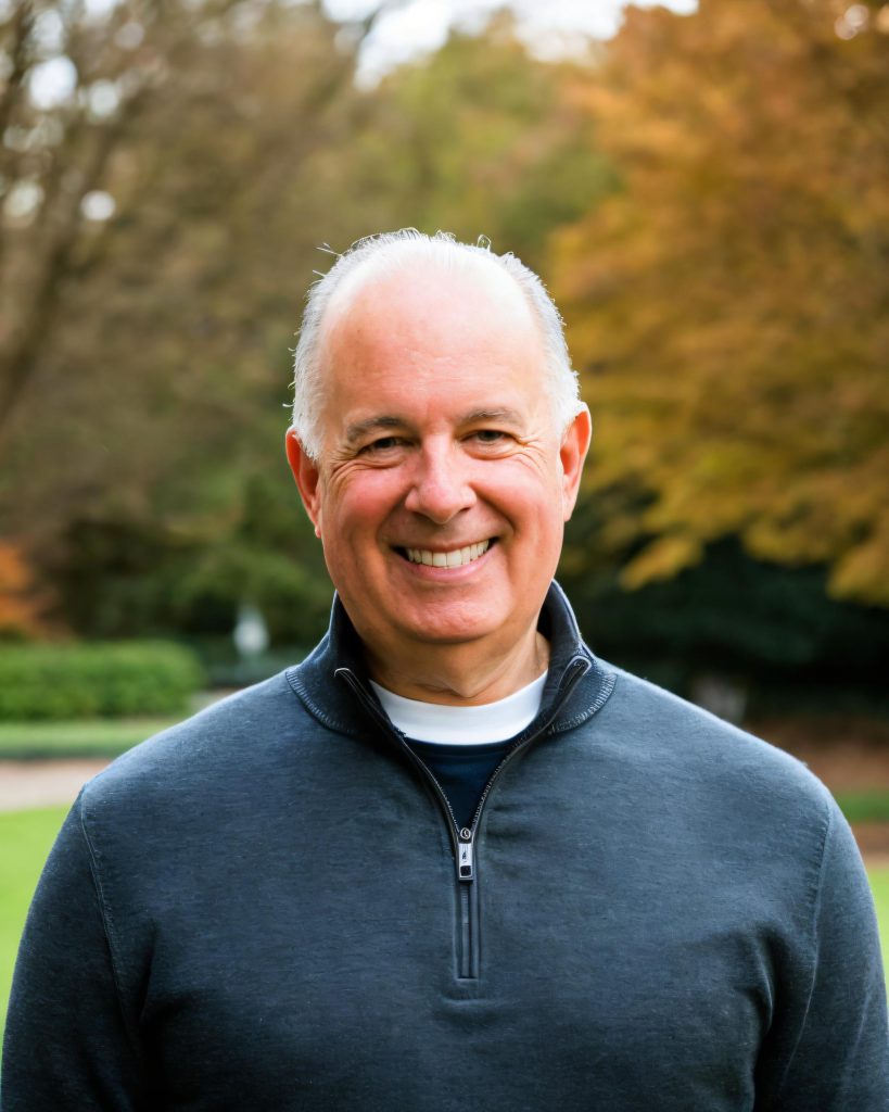 Person wearing a dark quarter-zip pullover over a white crew-neck shirt stands outdoors in a park setting with green grass and autumn trees in the background