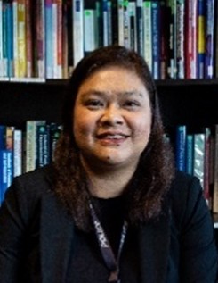 Person wearing a dark blazer and lanyard sits before tall bookshelves filled with assorted academic texts in a library or office setting.