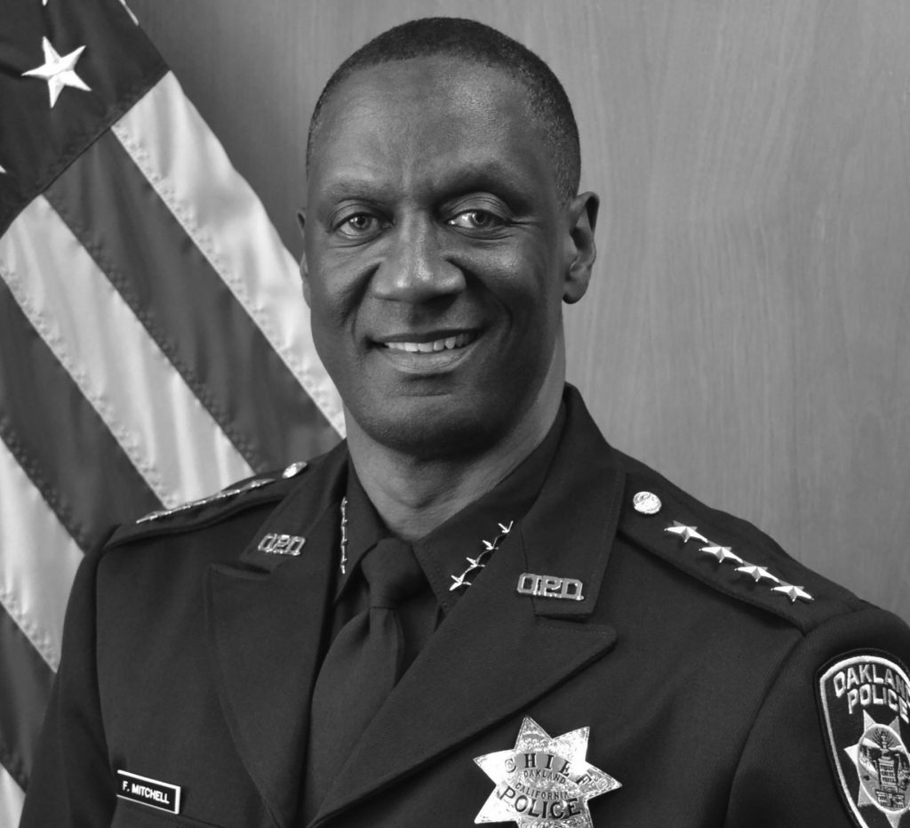 A black and white photograph of a uniformed police officer standing in front of an American flag. The officer is wearing a formal police uniform with a badge on the left chest that reads "POLICE" and has four stars on each shoulder epaulet, indicating a high rank. The uniform includes a tie and several pins or insignia on the collar. On the right shoulder, there is a patch that reads "OAKLAND POLICE." The background consists of a plain wall with the American flag prominently displayed to the left side of the image.