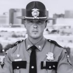 a black-and-white photograph of a law enforcement officer in uniform. The uniform includes a wide-brimmed hat with an emblem on the front, a buttoned shirt with shoulder epaulets, and various insignia and badges. One badge is prominently displayed on the left side of the chest, while another badge is pinned to the right side. A name tag is visible above the right pocket of the shirt. The officer also wears a tie and has two pins on each collar tab. In the background, there are blurred buildings and trees, indicating an urban setting.