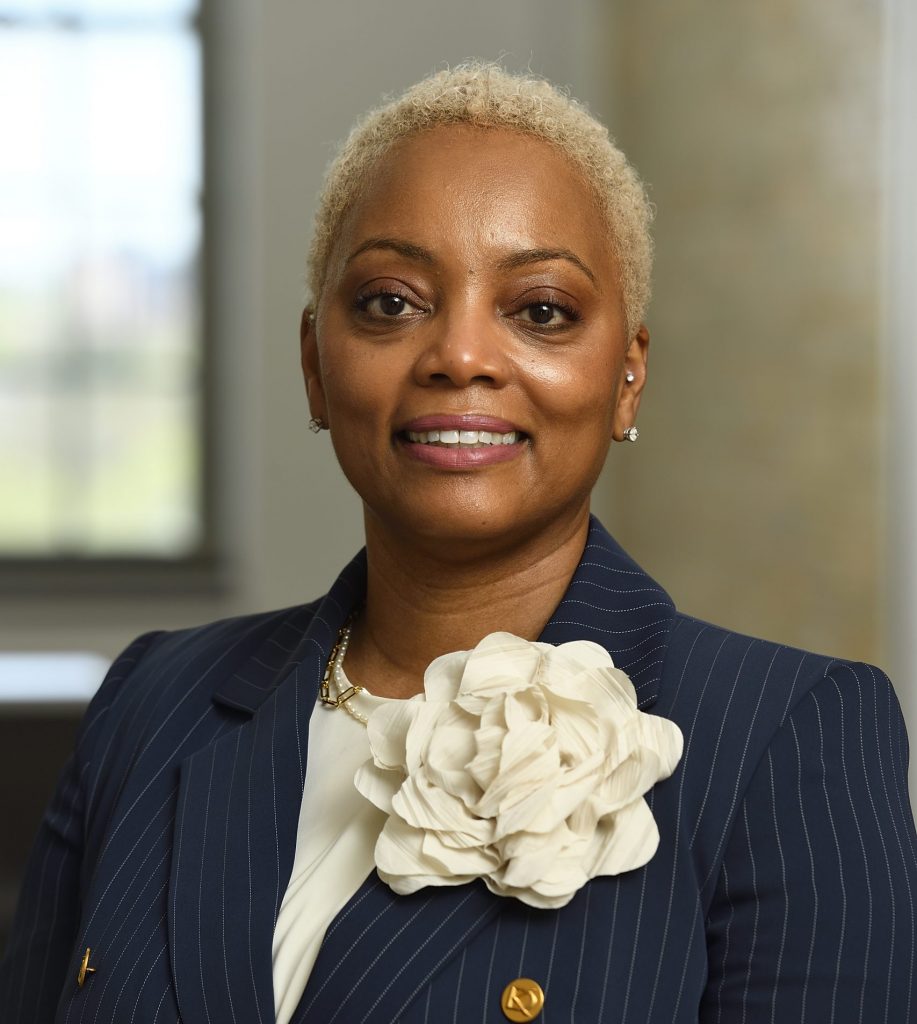 Person wearing a navy pinstripe blazer with gold buttons and a large white floral accessory on the lapel, paired with a white top, indoors near a window