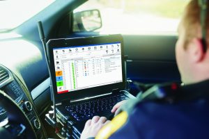 Police officer operates laptop in patrol car; screen shows task management software with icons, lists, and color-coded indicators for field operations