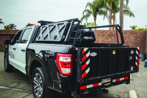 Black and white police pickup truck with modular cargo system, rear gate featuring red reflective stripes, mounted equipment, and palm trees in background