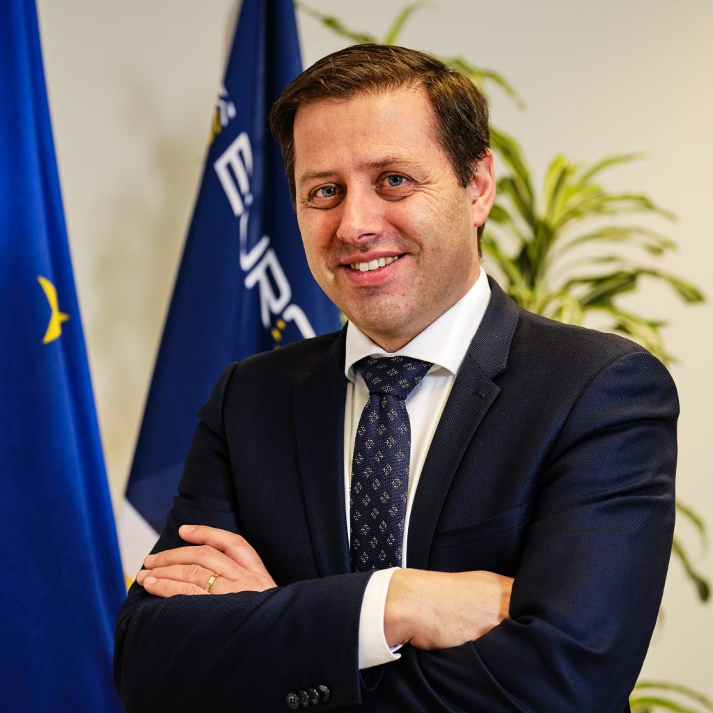 Individual in dark suit, white shirt, and patterned tie stands with arms crossed before blue and yellow flags; green plants visible in background.