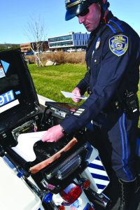 Uniformed motorcycle officer reviews papers beside an open police motorcycle case with emergency lights, radio gear and modern buildings behind