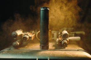 Black cylindrical suppressor upright on metallic surface, surrounded by four firearms aimed inward; smoky background adds dramatic effect.