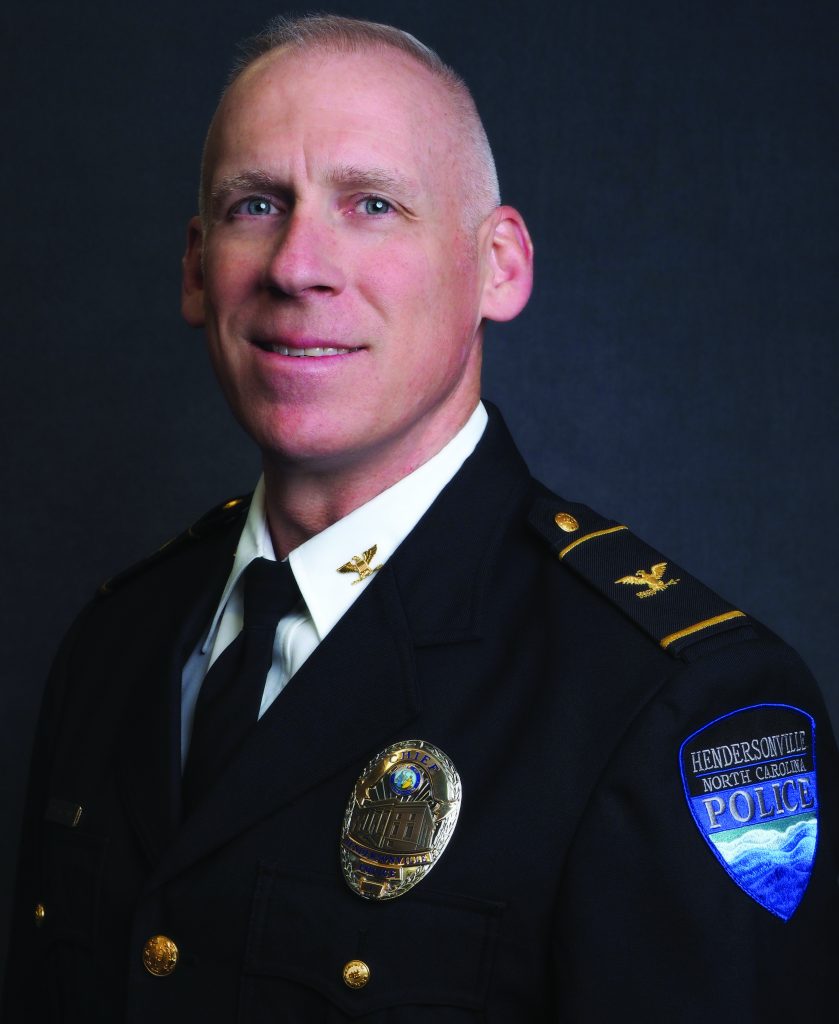 Police officer in formal dark uniform with gold buttons, badge, black tie, and shoulder patch reading Hendersonville North Carolina Police against gray backdrop