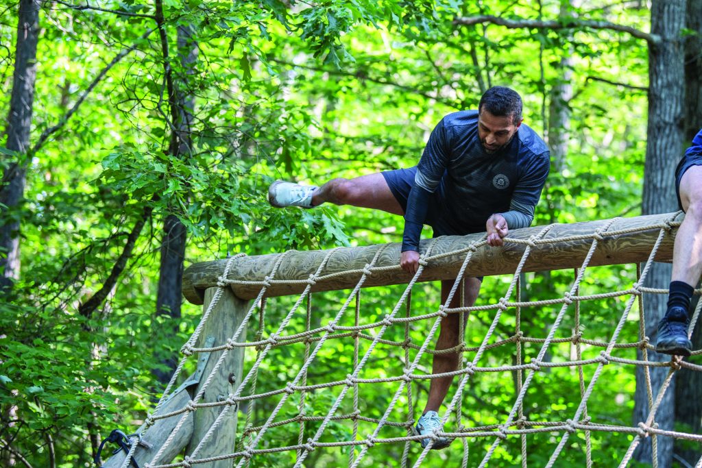 Participant in athletic wear climbs over a wooden obstacle with rope netting in a forest setting, gripping the top beam while stepping through the net structure