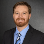 Professional studio portrait of an individual wearing a dark suit jacket, light blue dress shirt, and blue striped tie, posed against a textured gray backdrop with soft lighting and a head-and-shoulders composition