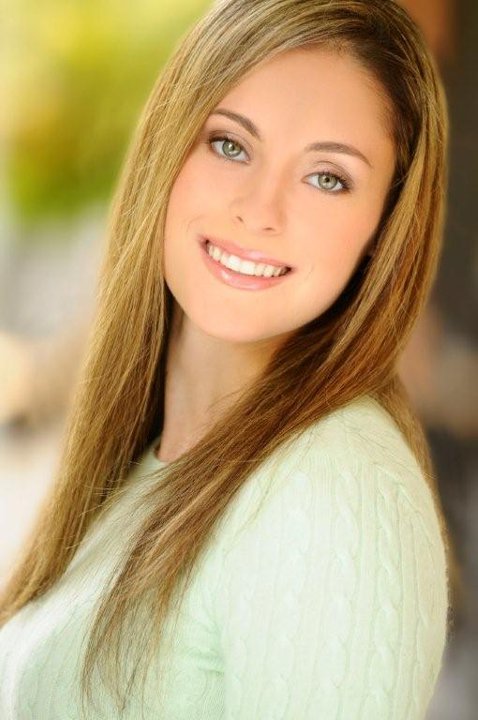 Portrait-style image of an individual wearing a light green sweater, shown from the shoulders up against a softly blurred outdoor background with greenery and warm natural light