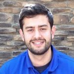 Person wearing blue collared shirt, standing before textured stone wall; image focuses on upper body and background surface detail