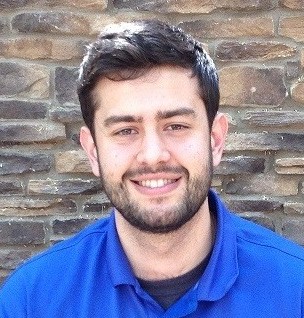 Person wearing blue collared shirt, standing before textured stone wall; image focuses on upper body and background surface detail