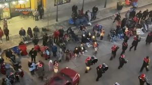 Overhead nighttime view of police and emergency responders surrounding multiple injured civilians lying on a city street and sidewalks, with onlookers, vehicles, drum carriers, and building storefronts illuminated by streetlights.